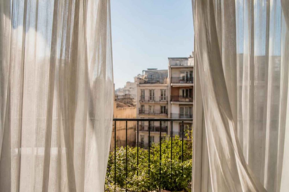 Widok na kamienicę i zielony balkon przez rozsunięte firany